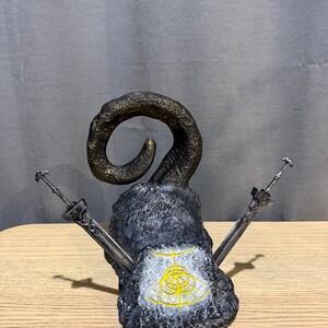 Op de afbeelding: Een gedetailleerde sculptuur met een grote, gebogen hoorn bovenop een getextureerde, grijze stenen basis. Twee zilveren zwaarden zijn aan weerszijden geplaatst. Een gouden embleem is zichtbaar op de voorkant van de basis. De sculptuur staat op een houten oppervlak.