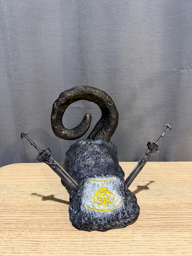Op de afbeelding: Een gedetailleerde sculptuur met een grote, gebogen hoorn bovenop een getextureerde, grijze stenen basis. Twee zilveren zwaarden zijn aan weerszijden geplaatst. Een gouden embleem is zichtbaar op de voorkant van de basis. De sculptuur staat op een houten oppervlak.
