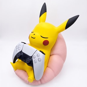 May include: A yellow Pikachu figure with black-tipped ears and red cheeks, sitting in a textured light pink chair. The figure is holding a white and black wireless video game controller in its hands, with its eyes closed in a relaxed expression.