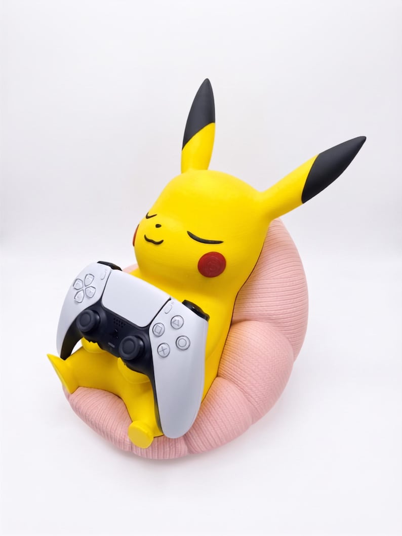 May include: A yellow Pikachu figure with black-tipped ears and red cheeks, sitting in a textured light pink chair. The figure is holding a white and black wireless video game controller in its hands, with its eyes closed in a relaxed expression.