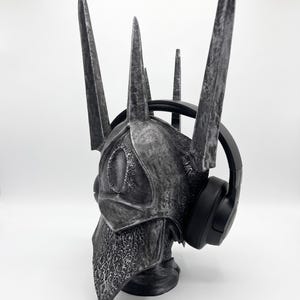 K&ouml;nnte beinhalten: Ein silber-schwarzer, verwitterter Helm mit einer hohen, stacheligen Krone und einem augenf&ouml;rmigen Emblem auf der Vorderseite, verziert mit filigranen Mustern am unteren Rand. Der Helm wird mit einem schwarzen Over-Ear-Kopfh&ouml;rer pr&auml;sentiert.