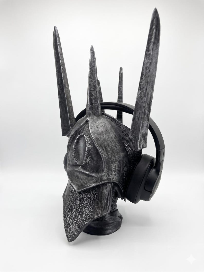K&ouml;nnte beinhalten: Ein silber-schwarzer, verwitterter Helm mit einer hohen, stacheligen Krone und einem augenf&ouml;rmigen Emblem auf der Vorderseite, verziert mit filigranen Mustern am unteren Rand. Der Helm wird mit einem schwarzen Over-Ear-Kopfh&ouml;rer pr&auml;sentiert.