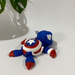 May include: A 3D-printed Captain America figurine in a lying-down pose. The figurine features a blue helmet with white wings, a red, white, and blue shield design on the chest, and red and blue limbs. The star on the shield is white.