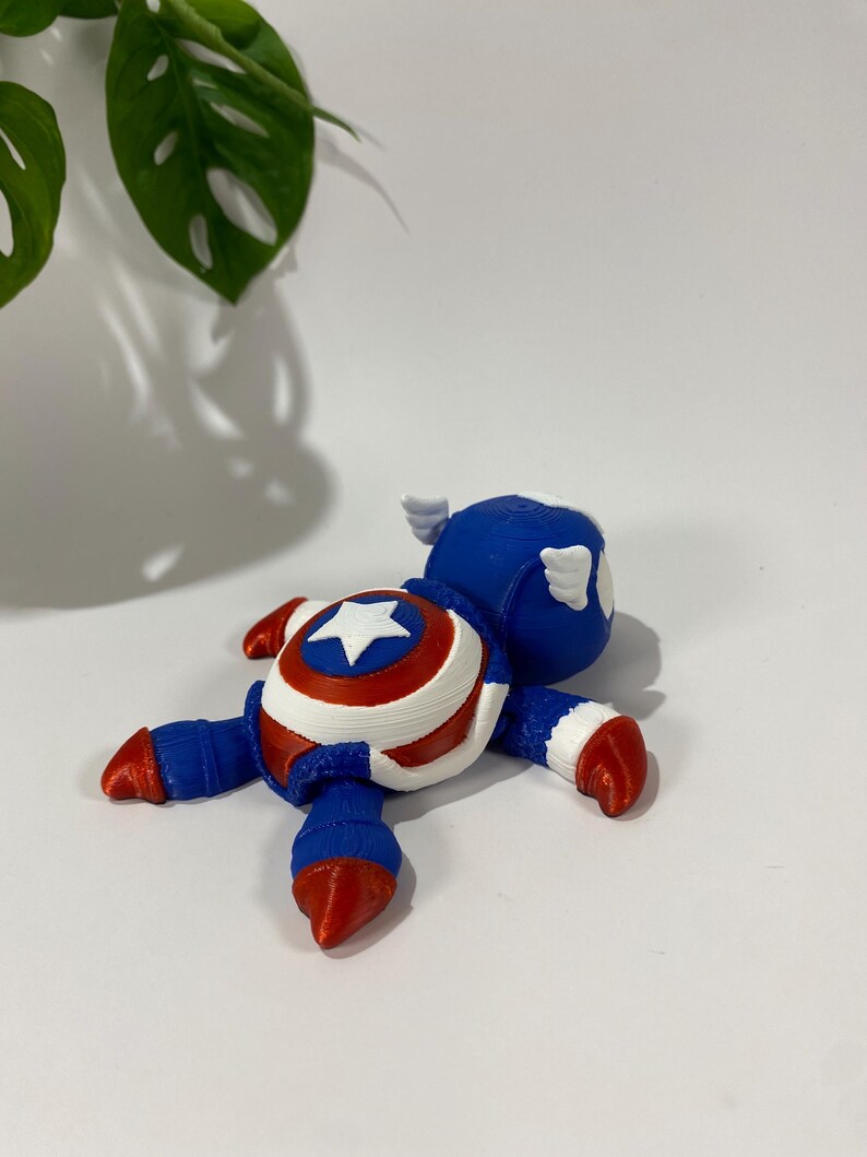 May include: A 3D-printed Captain America figurine in a lying-down pose. The figurine features a blue helmet with white wings, a red, white, and blue shield design on the chest, and red and blue limbs. The star on the shield is white.
