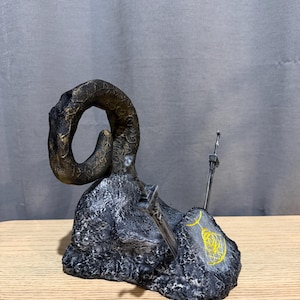 Op de afbeelding: Een gedetailleerde sculptuur met een grote, gebogen hoorn in bronskleuren, die oprijst uit een getextureerde, donkergrijze basis. Twee zilveren zwaarden zijn in de basis ingebed en een geel symbool is op de zijkant geschilderd. De sculptuur staat op een houten oppervlak.