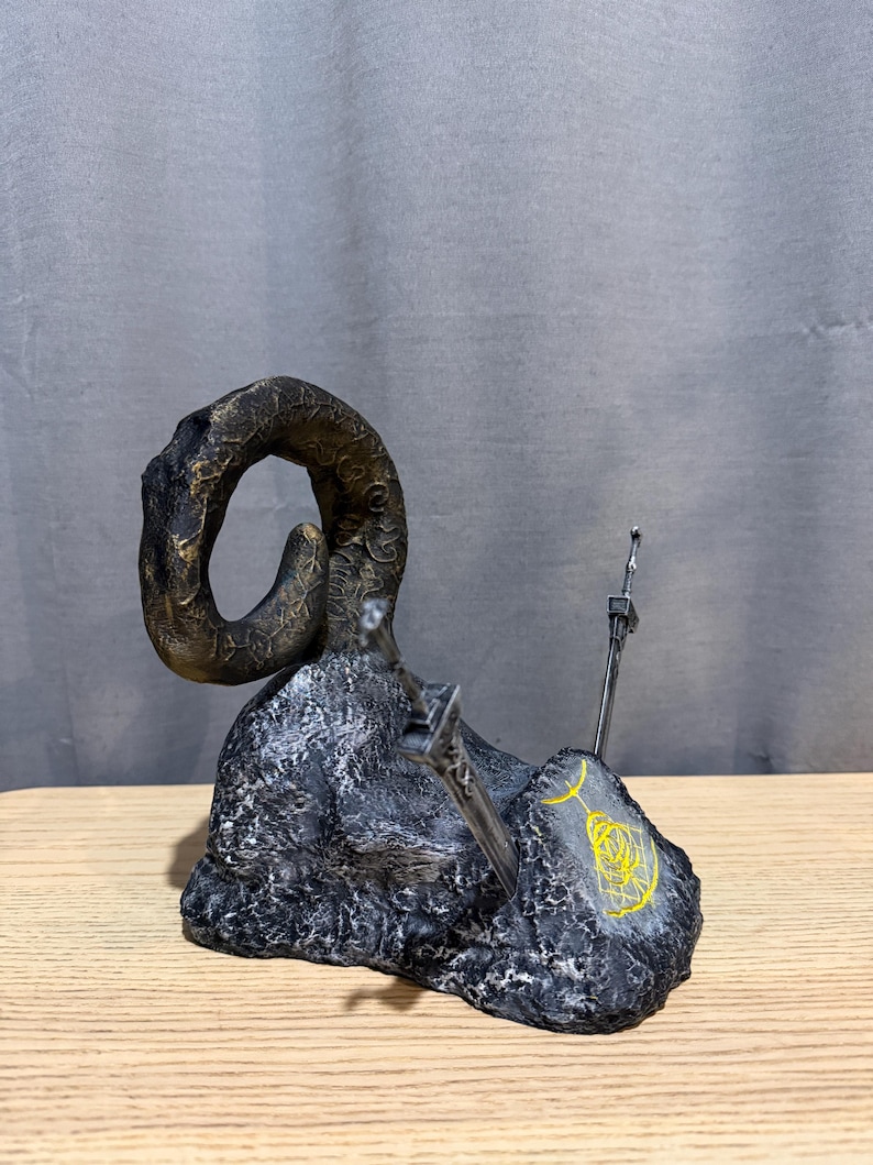Op de afbeelding: Een gedetailleerde sculptuur met een grote, gebogen hoorn in bronskleuren, die oprijst uit een getextureerde, donkergrijze basis. Twee zilveren zwaarden zijn in de basis ingebed en een geel symbool is op de zijkant geschilderd. De sculptuur staat op een houten oppervlak.