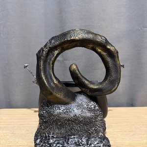 Op de afbeelding: Een bronzen sculptuur in de vorm van een gebogen hoorn, gemonteerd op een getextureerde, grijszwarte basis. De hoorn heeft een goudkleurige accent. De sculptuur staat tegen een neutrale achtergrond en staat op een houten oppervlak.