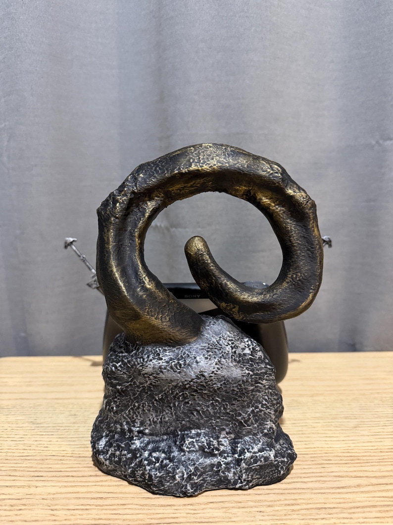 Op de afbeelding: Een bronzen sculptuur in de vorm van een gebogen hoorn, gemonteerd op een getextureerde, grijszwarte basis. De hoorn heeft een goudkleurige accent. De sculptuur staat tegen een neutrale achtergrond en staat op een houten oppervlak.