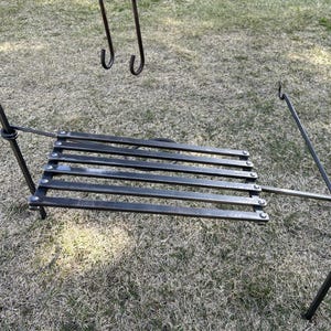 May include: A black metal campfire tripod with a grill grate and hanging hooks. The tripod has three legs and a horizontal grate for cooking. The hooks are for hanging pots or other cooking utensils. The tripod is set up on a grassy surface.