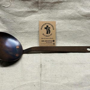 May include: A handcrafted, dark brown metal ladle with a rounded bowl and a long, flat handle. The bowl is riveted and has a dark, almost iridescent finish. The handle has a hole at the end for hanging. A business card is in the background.