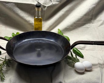 May include: A round, dark-colored metal skillet with a long handle and a small side loop handle. The pan is surrounded by two white eggs, two green jalapeño peppers, sprigs of fresh rosemary, and a small glass bottle filled with golden oil.