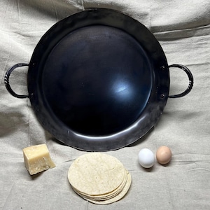 May include: A large, round, black cast iron pan with two handles. The pan is sitting on a beige fabric surface. There are two white eggs, one brown egg, a block of cheese, and a stack of tortillas next to the pan.