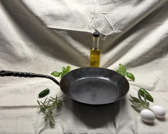 May include: A black carbon steel skillet with a long, twisted metal handle. The pan is surrounded by fresh basil leaves, rosemary sprigs, two green jalapeño peppers, two white eggs, and a small glass bottle filled with golden cooking oil.