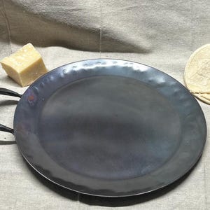 May include: A large, round, black metal serving tray with a hammered finish and a single handle. The tray is sitting on a beige fabric surface.