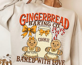 May include: A beige sweatshirt with a festive gingerbread theme. The design features the text "GINGERBREAD BAKING CO." in orange and red, along with gingerbread people, bows, a cookie jar, and candy canes. The phrase "BAKED WITH LOVE" is also included.