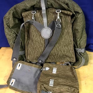 May include: A vintage olive green and brown camouflage backpack with multiple compartments and grey straps. The backpack features metal clasps and buckles, and a circular grey patch.