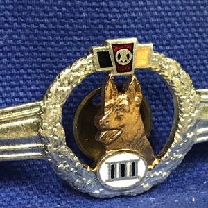 May include: A silver and gold pin featuring a dog silhouette in the centre. The dog is surrounded by a wreath of leaves and a white circle with the Roman numeral III. The pin has a silver wing on each side.