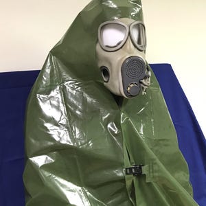 Op de afbeelding: Een groene plastic poncho met capuchon met een gasmasker bevestigd aan de capuchon. De poncho heeft een zwarte clip aan de zijkant.