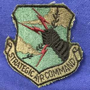 May include: Embroidered patch featuring the words "STRATEGIC AIR COMMAND" in gold lettering. The patch has a shield shape with a black and red design on a green background, and is bordered by a gold trim.