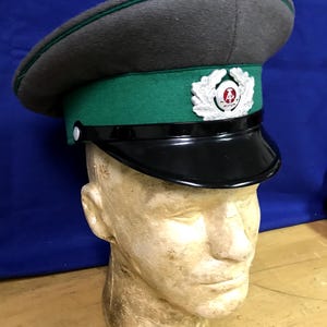 Könnte beinhalten: Eine graue Militärmütze mit grünem Band und schwarzem Visier. Die Mütze hat ein silbernes Emblem mit einem rot-weißen Design. Die Mütze wird auf einem Schaufensterkopf vor blauem Hintergrund präsentiert.