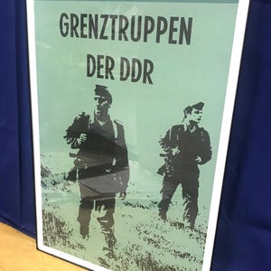 May include: A vintage poster with a green background and black text that reads "GRENZTRUPPEN DER DDR". Two soldiers in silhouette are walking in a field.