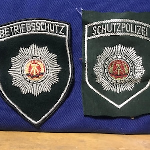 May include: Two dark green patches with white embroidered borders and text. One patch reads "BETRIEBSSCHUTZ" and the other reads "SCHUTZPOLIZEI". Both patches feature a central emblem with a red and gold design surrounded by a silver burst.