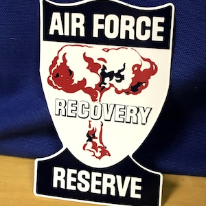 May include: A white, black, and red Air Force Recovery Reserve sticker. The sticker features the words "AIR FORCE" at the top, "RECOVERY" in the middle, and "RESERVE" at the bottom. A red and white graphic is in the center.