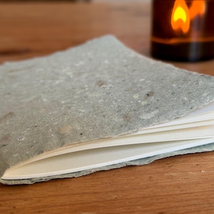 Könnte beinhalten: Ein kleines, graues, handgemachtes Notizbuch mit einem strukturierten Einband aus Recyclingpapier. Das Notizbuch ist geöffnet und zeigt leere weiße Seiten.