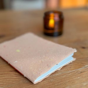 Könnte beinhalten: Ein kleines, rosafarbenes Notizbuch mit einem strukturierten Einband und weißen Seiten. Das Notizbuch liegt auf einem Holztisch neben einem kleinen, bernsteinfarbenen Glasgefäß.