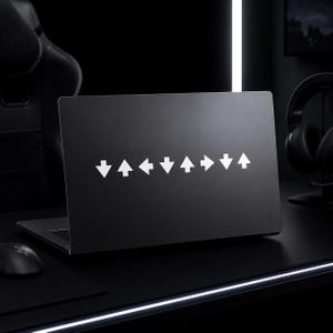 May include: Black laptop with a white arrow pattern on the lid. The arrows point up, down, left, and right, creating a directional design. The laptop is open on a black desk, with a mouse and headphones visible.