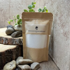 Puede incluir: Una bolsa de papel marr&oacute;n con una etiqueta blanca que dice "Vetiver and Teakwood Bath Salts 4 oz.". La bolsa est&aacute; llena de sales de ba&ntilde;o blancas y est&aacute; sobre una superficie de madera con piedras.