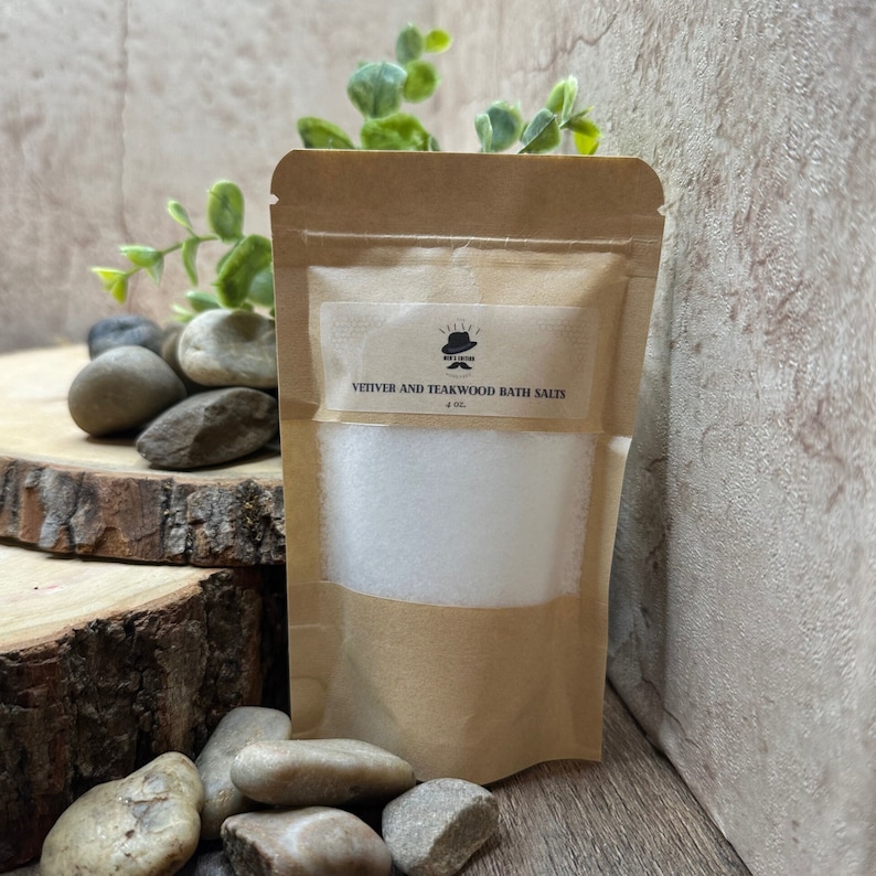 Puede incluir: Una bolsa de papel marr&oacute;n con una etiqueta blanca que dice "Vetiver and Teakwood Bath Salts 4 oz.". La bolsa est&aacute; llena de sales de ba&ntilde;o blancas y est&aacute; sobre una superficie de madera con piedras.