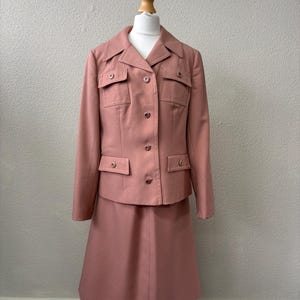 May include: A vintage rose-colored skirt suit displayed on a mannequin. The suit includes a collared jacket with four pockets and a matching knee-length skirt. The jacket has button closures. The suit is made of a solid, textured fabric.