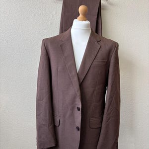 May include: A solid brown suit jacket featuring a notched lapel, two dark buttons on the front, and two flap pockets. The jacket is displayed on a mannequin, with matching brown trousers hanging on a wooden hanger positioned directly behind it.