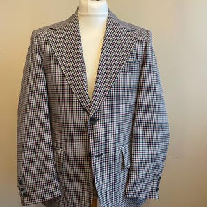 May include: A houndstooth patterned blazer on a mannequin. The jacket has a notched lapel, two front pockets, and buttoned cuffs. The colour scheme includes black, white, red, and green, creating a textured, checkered pattern. The blazer is a classic style.