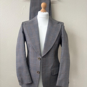May include: A grey blazer with a subtle brown and blue plaid pattern, featuring wide lapels, two front buttons, and two flap pockets. The blazer is displayed on a mannequin with matching grey plaid trousers hanging on a wooden hanger behind it.