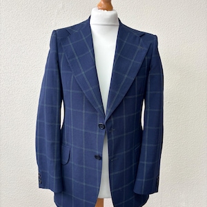 May include: A navy blue blazer with a plaid pattern in green and blue. The blazer has a two-button closure, notched lapels, and a chest pocket. The jacket is displayed on a wooden mannequin.