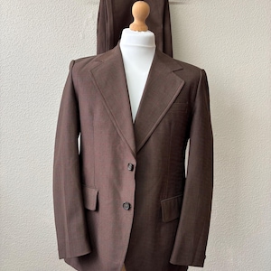 Puede incluir: Una americana marrón con un sutil patrón de cuadrícula oscuro, que presenta solapa de muesca, dos botones frontales y dos bolsillos con solapa. La americana se muestra en un maniquí, con pantalones marrones a juego colgados en una percha.