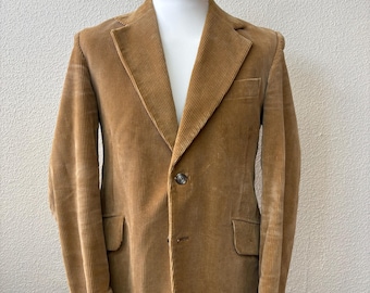 Pode incluir: Um blazer de veludo cotelê bege com lapela entalhada, fecho frontal de um botão, dois bolsos com aba na cintura e um bolso no peito. O casaco é exibido num manequim branco contra uma parede branca com textura.