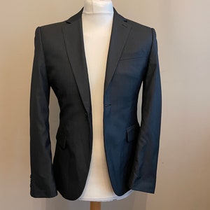 Può includere: Un blazer nero a righe sottili con una chiusura a un bottone. Il blazer è su un manichino di legno.