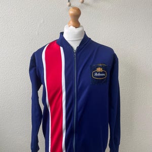 Può includere: Una giacca da tuta blu, rossa e bianca con chiusura a zip. La giacca ha una striscia rossa e bianca sul lato sinistro e una piccola toppa con il testo "Rothmans" sul petto destro.