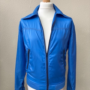 Op de afbeelding: Een felblauwe jas met een volledige ritssluiting, twee voorzakken en een geribde zoom. De jas heeft een klassieke kraag en lange mouwen. De jas wordt op een mannequin getoond.