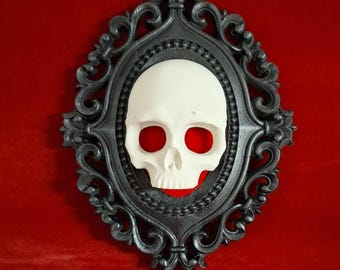 Op de afbeelding: Een decoratieve wanddecoratie met een witte schedel met rode oogkassen in een zwart, sierlijk frame. Het frame heeft een gedetailleerd krulwerkontwerp. De achtergrond is rood.