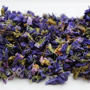 May include: Dried violet petals in shades of purple, blue, and green. The petals are loose and scattered on a white surface.