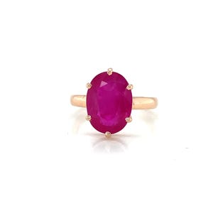 Könnte beinhalten: Ein goldener Ring mit einem großen, oval geschliffenen Rubin. Der Rubin hat einen leuchtenden rot-rosa Farbton, und der Ring hat ein schlichtes, elegantes Design. Der Ring ist vor einem weißen Hintergrund platziert.
