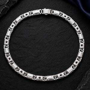 May include: A silver-toned chain necklace featuring a repeating pattern of rectangular links connected by circular rings. The metal has a polished, reflective finish, and the design includes a secure, integrated clasp mechanism at the top centre.