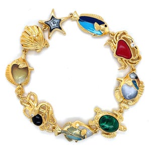 Könnte beinhalten: Goldfarbenes Armband mit Anhängern aus dem Meer. Das Armband enthält einen Seestern, eine Muschel, Fische, eine Krabbe, eine Schildkröte und einen Oktopus. Die Anhänger sind mit Emaille in verschiedenen Farben wie Blau, Rot, Grün und Schwarz detailliert.