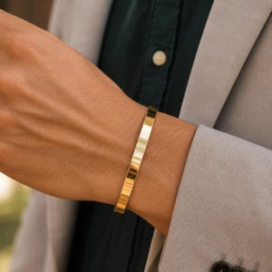 Könnte beinhalten: Ein poliertes Armband aus goldfarbenem Metall mit einer glatten, flachen Oberfläche und einem einfachen, offenen Design. Das Armband hat eine reflektierende Oberfläche, die das Licht einfängt und für ein klares Erscheinungsbild sorgt.