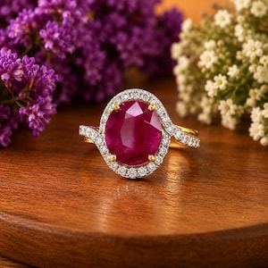 Op de afbeelding: Een gouden ring met een grote, ovaal geslepen, diep roze-rode edelsteen in het midden, omringd door een halo van kleine, heldere, rond geslepen stenen. De ringband is aan de zijkanten ook bezet met een rij kleine, heldere, rond geslepen stenen.