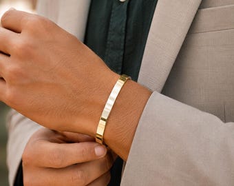 May include: A polished gold-tone cuff bracelet with a smooth, flat surface and a sleek, minimalist design. The bracelet features a continuous, solid band that wraps around the wrist, showcasing a reflective metallic finish and clean edges.